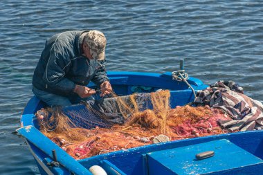 Bari, Puglia, Italy - April 8 2023: Fisherman with a hat repairing with a big needle a fishing net on an old blue wooden fishing boat at the port quay in Bari, Puglia Italy clipart