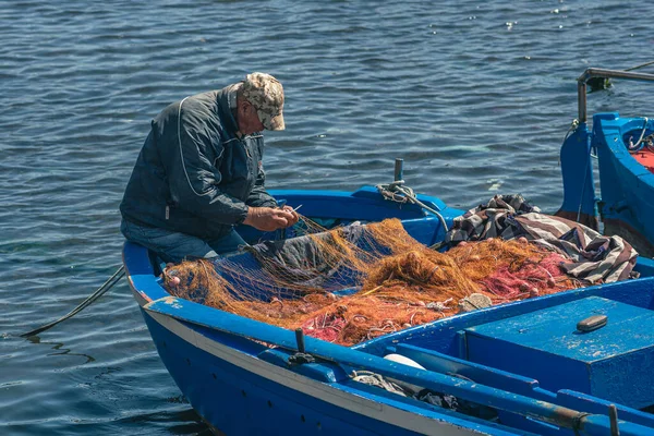 stock image Bari, Puglia, Italy - April 8 2023: Fisherman with a hat repairing with a big needle a fishing net on an old blue wooden fishing boat at the port quay in Bari, Puglia Italy