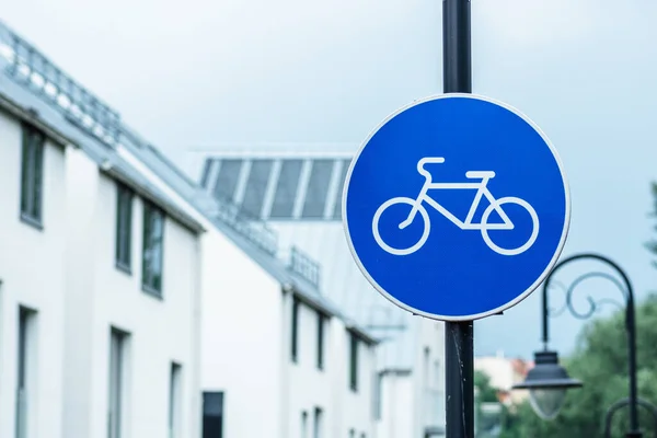 Blue road sign or signal of bicycle lane among new buildings and green trees, spring or summer nature and street lamps background, close up