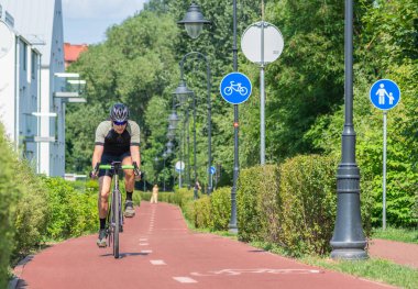 Vilnius, Litvanya - 4 Temmuz 2023: Yeşil ağaçlar ve çitler arasında bisiklet yolu işareti veya mavi yol işareti olan bisiklet yolunda bisiklet süren çocuk, ilkbahar yaz doğası ve sokak lambaları