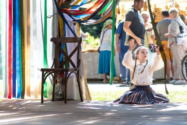 Riga, Letonya - 10 Temmuz 2023: Geleneksel giysiler içindeki güzel küçük kız XXVII. Letonya Şarkı ve XVII Dans Festivali sırasında sahnedeki boş bir sandalyenin yanında renkli çizgilerle oynuyor 