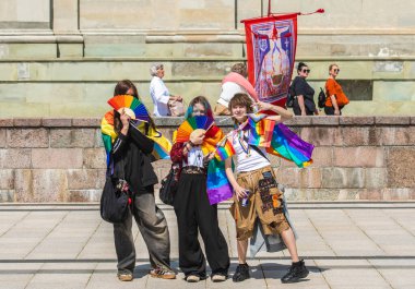 Vilnius, Litvanya - 7 Haziran 2025: Gökkuşağı renkli saçlı ve yelpazeli sevimli gençler, LGBT + gururu sırasında haç ve bayraklar takıyor ve arkasında Madonna 'nın resmiyle protesto yapan yaşlı bayanlarla birlikte