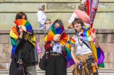 Vilnius, Litvanya - 7 Haziran 2025: Gökkuşağı renkli saçlı ve yelpazeli sevimli gençler, LGBT + gururu sırasında haç ve bayraklar takıyor ve arkasında Madonna 'nın resmiyle protesto yapan yaşlı bayanlarla birlikte