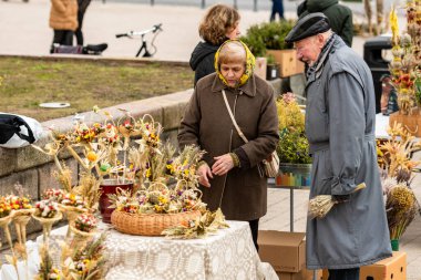 Vilnius, Litvanya - 13 Nisan 2025: Pazar günü Litvanya 'nın başkenti Vilnius' ta bulunan katedralin yakınındaki kurutulmuş çiçek ve bitkilerden yapılan geleneksel el işi süslemeler olan verbo satan yaşlı kadın