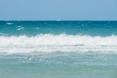 Okyanus dalgalarının, ufukta görünen bir yelkenliyle berrak mavi gökyüzünün altında kıyıya çarptığı manzara. Sakin bir deniz manzarası doğanın ve açık denizin güzelliğini yansıtıyor.