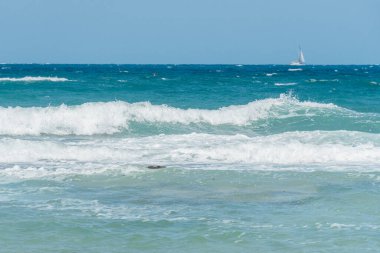 Okyanus dalgalarının, ufukta görünen bir yelkenliyle berrak mavi gökyüzünün altında kıyıya çarptığı manzara. Sakin bir deniz manzarası doğanın ve açık denizin güzelliğini yansıtıyor.