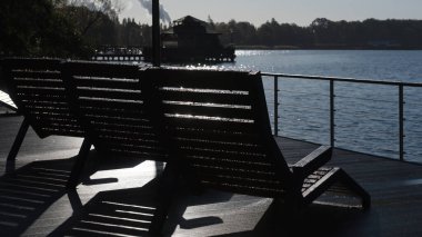 LANDSCAPE BY LAKE - Morning sun over the wooden lounge chairs in the drops of morning dew on the viewing platform clipart