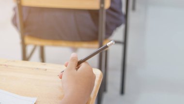 Sınav kâğıdı yazmak için ellerinde kalem tutan Asyalı öğrencilerin bulanıklığı. Sınav odasında öğrenci üniforması ve eğitim konsepti olan tahta masa masasına sınav kağıtları.
