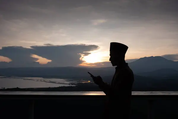 Silhouette of an Asian Muslim man holding a smartphone under the sunset. A man holding a smartphone under the cloudy sunset.