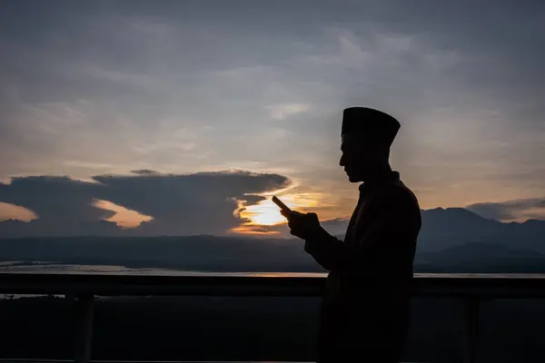 Silhouette of an Asian Muslim man holding a smartphone under the sunset. A man holding a smartphone under the cloudy sunset.