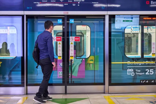 Mavi gömlekli ve siyah pantolonlu bir adam tren istasyonunda yürüyor. Tren peronda ve adam bir sırt çantası taşıyor.