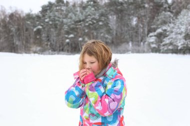 A child girl in winter clothes makes a snowman, rolls big snowballs, sits on a snowball, blows on hands to warm up. Winter entertainment, activities and leisure. Snowy forest and field, snowfall