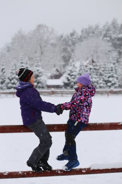 Two smiling laughing girls sisters siblings sitting on farm fence under snowfall. Farm in the pine fir tree forest.Witer activities sports and leisure