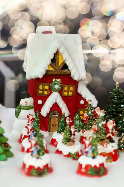 Red toy house surrounded Christmas trees and Santas. Home decor on a background of snow and bokeh New Year festive glowing decoration. Concept of cozy home in winter.