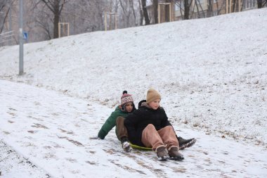Çocuklar karlı bir parkta yürüyor, yürüyor, kızakla kayıyor ve oynuyorlar. Çocuklarla kış eğlencesi ve açık hava aktiviteleri