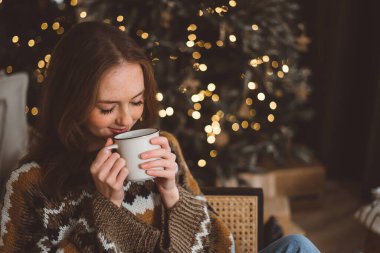 Çekici bir genç kadının portresi. Sıcak bir süveter giymiş ve elinde kırmızı bir kupayla bir Noel ağacının arka planında. Kız rahat bir Yeni Yıl arifesinde içki içiyor..