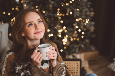Hoş, kızıl saçlı bir kadın, bir Noel ağacının arka planında, sıcak bir oturma odasında, elinde bir fincan çayla. Neşeli kızıl saçlı bayan topu Noel ağacına koyuyor. Evde kadın hazırlığı