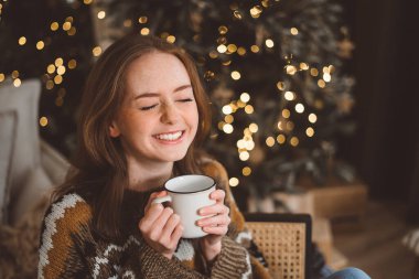 Hoş, kızıl saçlı bir kadın, bir Noel ağacının arka planında, sıcak bir oturma odasında, elinde bir fincan çayla. Neşeli kızıl saçlı bayan topu Noel ağacına koyuyor. Evde kadın hazırlığı
