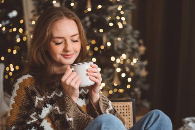 Çekici bir genç kadının portresi. Sıcak bir süveter giymiş ve elinde kırmızı bir kupayla bir Noel ağacının arka planında. Kız rahat bir Yeni Yıl arifesinde içki içiyor..