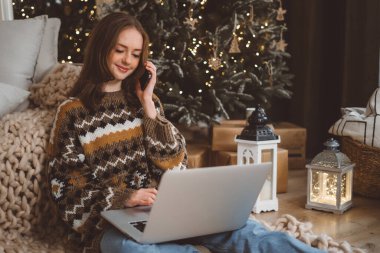 Sade süveterli, kızıl saçlı genç bir kadın, evinde Noel ağacı olan aile ve arkadaşlarıyla cep telefonuyla yaptığı telefon görüşmesini kullanıyor. Yüksek kalite fotoğraf