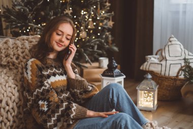 Hoş bir oturma odasındaki bir Noel ağacının arka planında oturan genç, çekici kızıl saçlı bir kadın cep telefonunu tutuyor ve video aracılığıyla ailesiyle el sallıyor. Yüksek kalite fotoğraf