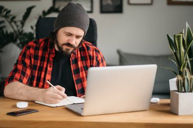 Odaklanmış sakallı genç adam, günlük giysiler içinde dizüstü bilgisayar kullanıyor, klavyeye yazıyor, e-posta ya da mesaj yazıyor, sohbet ediyor, alışveriş yapıyor, bilgisayar üzerinde çalışan başarılı bir serbest yazar. 
