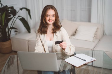 Neşeli genç kızıl saçlı kadın online alışveriş için akıllı telefon alışveriş uygulaması kullanıyor, kredi kartı tutuyor ve banka bilgilerini giriyor, evdeki oturma odasındaki sıradan atmosferde cam masada oturuyor.