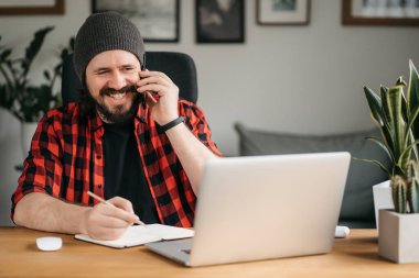 Mutlu, modern beyaz adam, dizüstü bilgisayar kullanırken cep telefonuyla konuşuyor, evde oturuyor, arkadaşlarıyla veya meslektaşlarıyla hoş sohbetler ediyor, yüzünü çeviriyor, gülümsüyor. Yüksek kalite fotoğraf