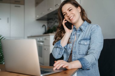 Genç bir kadın mutfak masasında dizüstü bilgisayar kullanarak oturuyor ve cep telefonuyla konuşuyor. Yüksek kalite fotoğraf