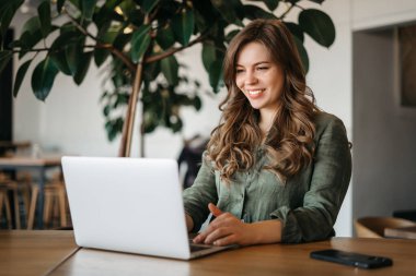 Rahat bir kafede masa başında otururken bilgisayarını uzaktan kontrol etmek için kullanan kendine güvenen bir kadın. Akıllı genç bir kadın e-postaları cevaplıyor, web sörfü yapıyor, kapalı mekanda, yan görüntüde.