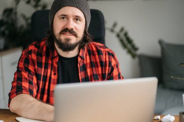 Masada dizüstü bilgisayarın önünde sakallı bir adam oturuyor. Şapka takıyor ve sakalı ekose desenle uyuşuyor.