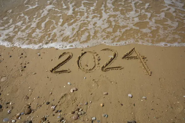 New Year 2024, lettering on the beach with wave and clear blue sea. Numbers 2024 year on the sea shore, message handwritten in the golden sand on beautiful beach background. New Years concept.