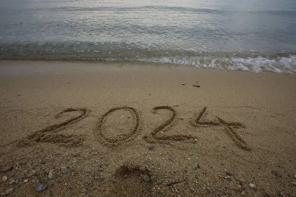 New Year 2024, lettering on the beach with wave and clear blue sea. Numbers 2024 year on the sea shore, message handwritten in the golden sand on beautiful beach background. New Years concept.