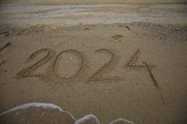 Yeni yıl 2024, plajda dalgalar ve açık mavi denizle yazılar. Deniz kıyısında 2024 numara, güzel plaj arka planında altın kumda el yazısıyla yazılmış mesaj. Yeni yıl kavramı.