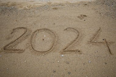 Yeni yıl 2024, plajda dalgalar ve açık mavi denizle yazılar. Deniz kıyısında 2024 numara, güzel plaj arka planında altın kumda el yazısıyla yazılmış mesaj. Yeni yıl kavramı.