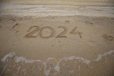 Yeni yıl 2024, plajda dalgalar ve açık mavi denizle yazılar. Deniz kıyısında 2024 numara, güzel plaj arka planında altın kumda el yazısıyla yazılmış mesaj. Yeni yıl kavramı.