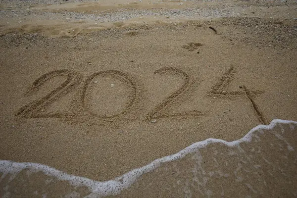 stock image New Year 2024, lettering on the beach with wave and clear blue sea. Numbers 2024 year on the sea shore, message handwritten in the golden sand on beautiful beach background. New Years concept.