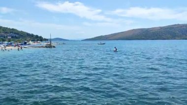 Bağlı bir yata yaklaşan küçük bir teknenin görüntüsü, arka planda deniz yüzüyor, insanlar denizde gülüp eğleniyor.