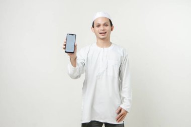 Gülümseyen Asyalı genç Müslüman beyaz arka planda izole bir şekilde cep telefonu ekranı gösteriyor. İnsanlar dini İslam yaşam tarzı kavramını benimsiyor. Kutlama Ramazan ve med Mübarek