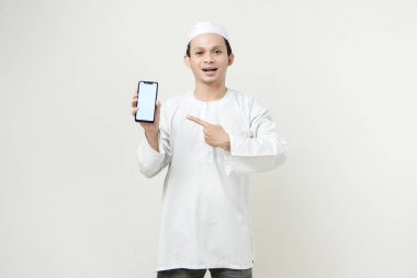 Mutlu Asyalı Müslüman boş telefon ekranı gösteriyor ve işaret işareti yapıyor. İnsanlar dini İslam yaşam tarzı kavramını benimsiyor. Ramazan ve Mübarek bayramını kutluyoruz. izole edilmiş arka grupta