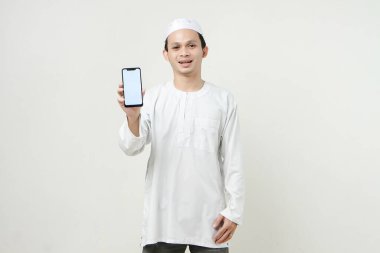 Gülümseyen Asyalı genç Müslüman beyaz arka planda izole bir şekilde cep telefonu ekranı gösteriyor. İnsanlar dini İslam yaşam tarzı kavramını benimsiyor. Kutlama Ramazan ve med Mübarek