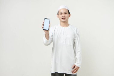 Gülümseyen Asyalı genç Müslüman beyaz arka planda izole bir şekilde cep telefonu ekranı gösteriyor. İnsanlar dini İslam yaşam tarzı kavramını benimsiyor. Kutlama Ramazan ve med Mübarek