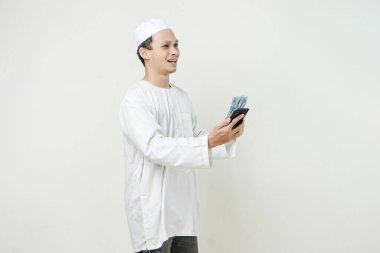 Rupiah banknotları ve cep telefonu elinde tutan mutlu Müslüman adam. İnsanlar dini İslam yaşam tarzı kavramını benimsiyor. Ramazan ve Mübarek bayramını kutluyoruz. izole edilmiş arka grupta