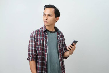 Düşünceli Asyalı adam izole edilmiş bir arka planda akıllı telefon düşünüyor ve tutuyor.