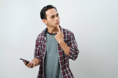 Düşünceli Asyalı adam izole edilmiş bir arka planda akıllı telefon düşünüyor ve tutuyor.