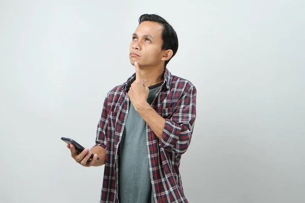 pensive asian man thinking and holding smartphone on isolated background