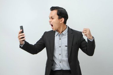 Kızgın Asyalı takım elbiseli iş adamı izole edilmiş arka planda akıllı telefon tutuyor.
