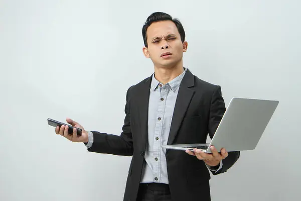 Angry mad asian indonesian business man in suit holding smart phone and laptop computer on isolated background