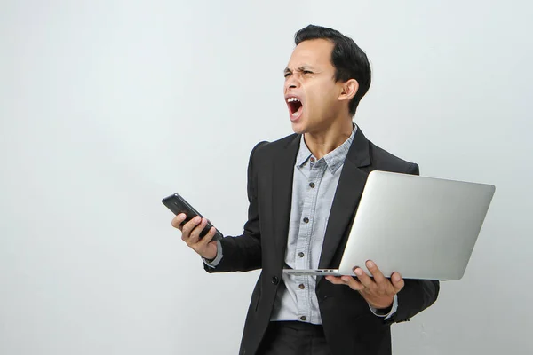 Angry mad asian indonesian business man in suit holding smart phone and laptop computer on isolated background