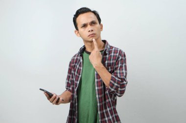 Düşünceli Asyalı adam izole edilmiş bir arka planda akıllı telefon düşünüyor ve tutuyor.